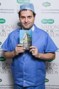 Photograph of Adam Kay wearing blue scrubs holding his book "This Is Going to Hurt" at the National Book Awards